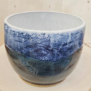 May include: A handcrafted ceramic bowl with a distinctive layered design. The bowl showcases a gradient of colours, shifting from a light, speckled white at the rim to a deep, dark blue at the base. The bowl's interior is a clean white, contrasting with the textured exterior.