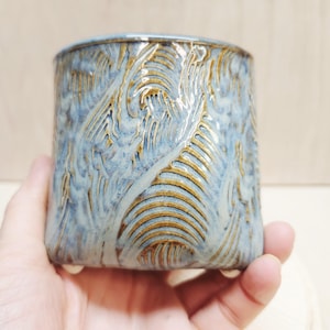 May include: A handmade ceramic tumbler with a textured, swirling design. The tumbler features a blend of blue and brown glazes, creating a unique, organic pattern. The cylindrical shape has a slightly flared base. The tumbler is held in a hand.