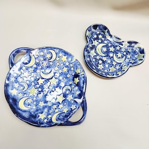 May include: Two ceramic dishes with a celestial theme. One is round with handles, the other is an irregular shape. Both are painted with a dark blue background, yellow stars, and crescent moons. The dishes are handmade.