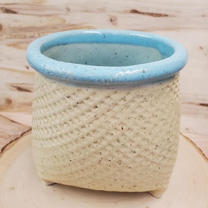 May include: A white ceramic planter with a light blue rim and a woven pattern. The planter has three small feet.
