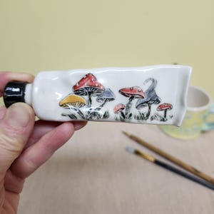 May include: A white ceramic tube-shaped container with a black cap, decorated with colorful mushrooms. The mushrooms have red, yellow, and gray caps. The container is held in a person's hand.