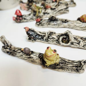 May include: Hand-painted ceramic log ornaments with forest creatures. Each log features a unique design, including a frog, mushrooms, and a snail. The logs are painted in naturalistic colours, with detailed textures.