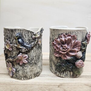May include: Two cylindrical ceramic mugs with a textured bark-like finish. The left mug features a blue bird perched on a branch with pink flowers. The right mug displays a large, detailed pink flower with green leaves and small buds.