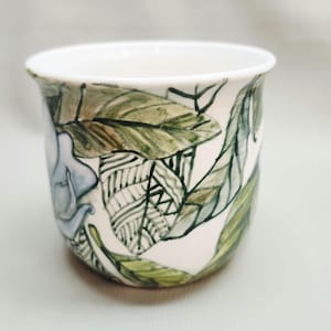 May include: A white ceramic mug featuring a hand-painted botanical design with green leaves and a light blue flower. The artwork includes detailed line work and shading, with some leaves displaying intricate geometric patterns in black ink.