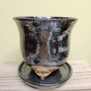May include: A handcrafted ceramic planter with a dark, glossy finish and a matching saucer. The planter has a unique, textured surface with a flared rim and sits on a small, decorative base. The saucer has a raised edge.