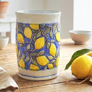 Peut inclure: Vase en céramique avec un fond bleu et un motif de citrons jaunes et de feuilles vertes. Le vase a un bord et une base blancs avec une bande bleue. Le design est en relief, lui donnant un aspect texturé.