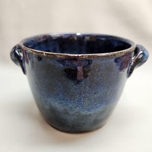 May include: A handmade ceramic bowl with a dark blue glaze. The bowl has two small handles and a slightly flared rim. The glaze has a mottled appearance, with lighter blue and brown accents. The bowl is suitable for serving soups or stews.