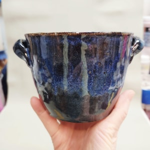 May include: A handcrafted ceramic bowl with a dark blue and black glaze. The bowl features two small handles and a textured surface. The rim is a darker brown colour. The bowl is held in a person's hand.