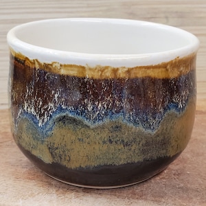 May include: A ceramic bowl with a white interior and a textured exterior. The exterior features bands of brown, gold, blue, and green hues. The bowl has a rounded shape and a slightly angled rim.