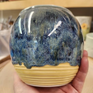 May include: A handcrafted ceramic vase with a rounded shape. The top half is glazed in shades of blue and green, while the bottom features a textured, horizontal yellow band. The vase is held in a hand, showcasing its size and design.