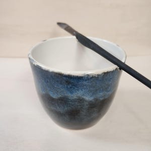 May include: A ceramic bowl with a gradient of blue and black hues, featuring a white interior and a textured rim. A black tool rests across the bowl's opening. The bowl's design suggests it is handmade.