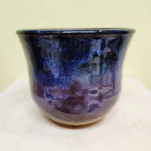 Peut inclure: Une tasse en céramique faite à la main avec un bord évasé. La tasse présente un glaçage brillant avec un mélange de teintes bleu foncé et violet. Les couleurs créent un effet marbré, ajoutant un intérêt visuel. Idéale pour les boissons.