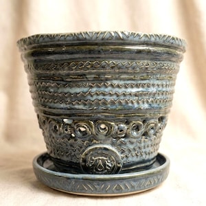 May include: A blue and grey ceramic planter with a matching saucer. The pot features a flared rim, intricate horizontal bands of zigzag and geometric textures, and a row of circular cutouts above a central circular medallion emblem.