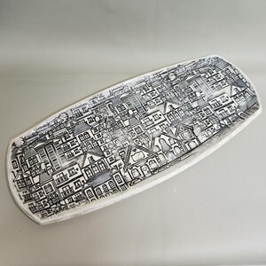 May include: A white ceramic serving platter with a detailed black and white illustration of a cityscape. The artwork depicts buildings, houses, and structures, creating a textured, artistic design.