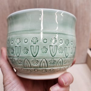 May include: A handmade, pale green ceramic bowl with a white base. The bowl has a repeating pattern of stylized shapes, including flowers, arches, and the letter "V". The bowl is held in a hand.