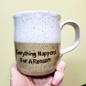 May include: A speckled ceramic mug with a white top and a tan bottom. The mug has a curved handle and the words "Everything Happens For A Reason" are printed on the front in black.