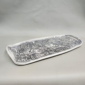 May include: A rectangular serving tray with a white border and a black and white cityscape design. The tray has a handle on one end. The tray is suitable for serving food or displaying decorative items.