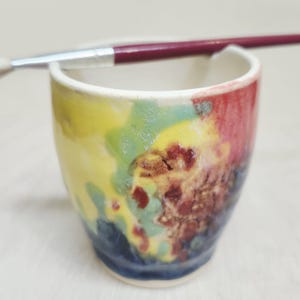 May include: A small ceramic cup with a smooth, rounded rim and a multi-colored glaze featuring abstract patches of yellow, green, red, and dark blue. A thin paintbrush with a dark red handle rests horizontally across the top of the cup rim.