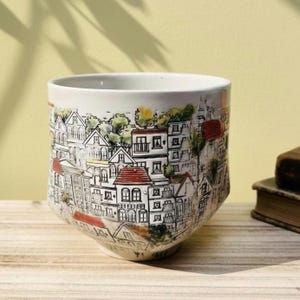 May include: White ceramic mug featuring a detailed townscape illustration. The design includes buildings with red, white, and grey roofs, and black outlines. The mug has a unique, tapered shape, suitable for hot drinks.