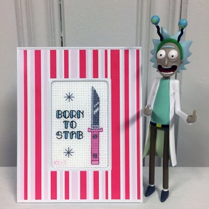 May include: A white frame with pink and red stripes surrounds a cross-stitch design. The design features a pink and black knife with the text "Born to Stab" above it. A cartoon figure of Rick Sanchez from the show Rick and Morty is standing to the right of the frame.
