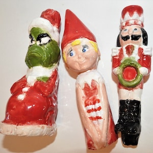 May include: Three ceramic Christmas ornaments: a green Grinch with a red Santa suit, a blonde elf with a red and white striped dress, and a red and black nutcracker holding a green wreath.