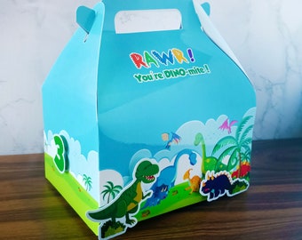 Puede incluir: Una caja de regalo para fiestas de color azul y verde con temática de dinosaurios y con asa. La caja tiene las palabras "RAWR! You're DINO-mite!" impresas en la parte delantera. La caja está decorada con dinosaurios y palmeras.
