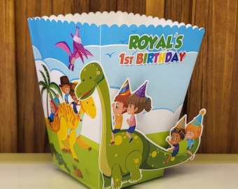 Puede incluir: Una caja de regalo de fiesta colorida con un dinosaurio y niños montando en él. La caja tiene un fondo azul con el texto "Royal's 1st Birthday" impreso en ella.