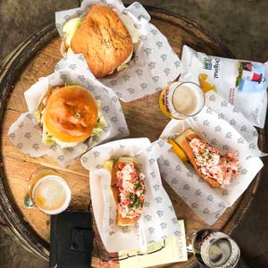 May include: A wooden table with four white paper containers holding various food items, including a lobster roll, a burger, and a sandwich. Two glasses of beer and a bag of potato chips are also on the table. The bag of chips is labeled "Original" and features a lighthouse.