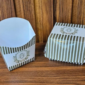 Puede incluir: Dos recipientes de papel para alimentos. Uno es abierto y en forma de cono, el otro es una caja cerrada. Ambos tienen un fondo blanco con rayas verticales verde oliva y un emblema decorativo.