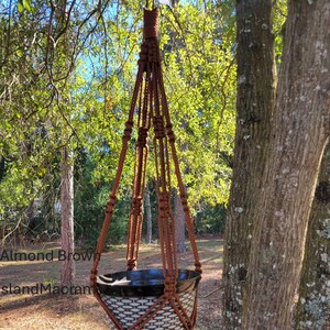 May include: Almond brown macrame plant hanger with a woven basket. The plant holder is suspended from a tree branch. The text "Almond Brown" and "Island Macramé" are visible on the image. The background features trees and sunlight.