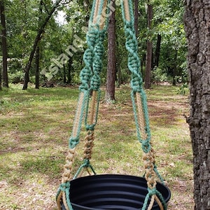 May include: A macrame plant hanger made with natural jute and green cotton rope. The hanger is holding a blue plastic planter. The hanger is hanging from a tree branch in a wooded area.