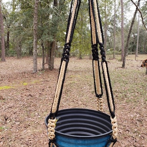 May include: A black and beige macrame plant hanger with a blue plastic pot hanging from it. The hanger is made with thick rope and has a knot pattern. The pot is empty and has a ribbed texture. The hanger is hanging in a wooded area.