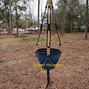 May include: A black and tan macrame plant hanger with a blue plastic pot hanging from a tree branch. The text "Island Macrame" is visible below the pot.