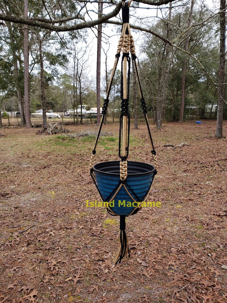 May include: A black and tan macrame plant hanger with a blue plastic pot hanging from a tree branch. The text "Island Macrame" is visible below the pot.