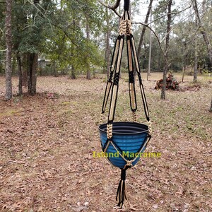 May include: A black and beige macrame plant hanger with a blue plastic pot hanging from it. The hanger is suspended from a tree branch in a wooded area.