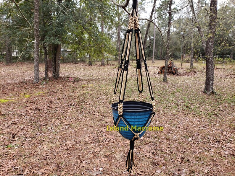 May include: A black and beige macrame plant hanger with a blue plastic pot hanging from it. The hanger is suspended from a tree branch in a wooded area.