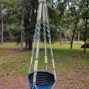 May include: A macrame plant hanger with a blue planter. The hanger is made with white and green rope and has a knotted design. The planter is hanging from a tree branch.