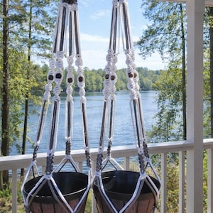 May include: Two macrame hanging planters with gray, white, and brown accents. The planters are suspended from a porch railing with a lake and trees in the background. The text "Island Macrame Gray & White" is visible.