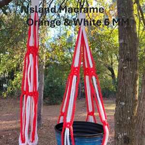 May include: Macrame plant hanger in orange and white, suspended from a tree branch. The text "Island Macrame Orange & White 6 MM" is visible. The hanger is designed to hold a blue pot, showcasing a handcrafted, decorative item.