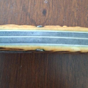 Puede incluir: Primer plano del mango de un cuchillo antiguo. El mango es de color marr&oacute;n claro y beige con un aspecto texturizado. La hoja es plateada con tres l&iacute;neas paralelas. El cuchillo descansa sobre una superficie de madera.