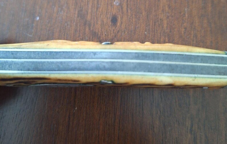 Puede incluir: Primer plano del mango de un cuchillo antiguo. El mango es de color marr&oacute;n claro y beige con un aspecto texturizado. La hoja es plateada con tres l&iacute;neas paralelas. El cuchillo descansa sobre una superficie de madera.