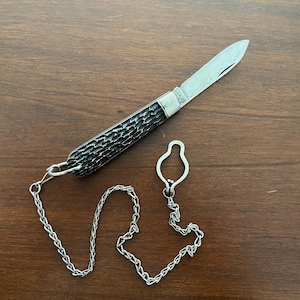 May include: A pocket knife with a textured black handle and a silver blade. The knife is attached to a silver chain and a key ring-style clasp. The knife is lying on a wooden surface.