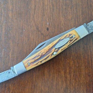 Puede incluir: Una navaja de bolsillo Case vintage con dos hojas. El mango tiene un patr&oacute;n amarillo y marr&oacute;n, con un logotipo Case plateado. Las hojas muestran signos de desgaste. La navaja est&aacute; sobre una superficie de madera.