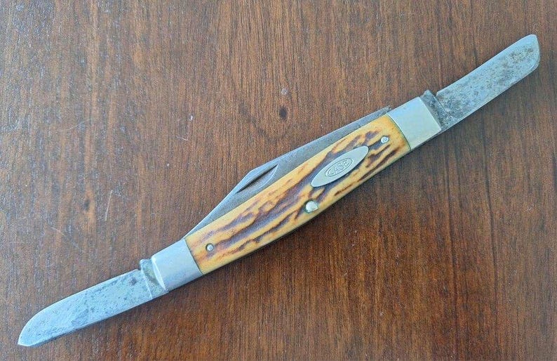 Puede incluir: Una navaja de bolsillo Case vintage con dos hojas. El mango tiene un patr&oacute;n amarillo y marr&oacute;n, con un logotipo Case plateado. Las hojas muestran signos de desgaste. La navaja est&aacute; sobre una superficie de madera.