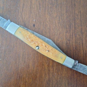 Puede incluir: Una navaja de bolsillo vintage con dos hojas. La navaja presenta un mango de madera de color claro con refuerzos y remaches met&aacute;licos. Las hojas son de acero y muestran signos de desgaste.