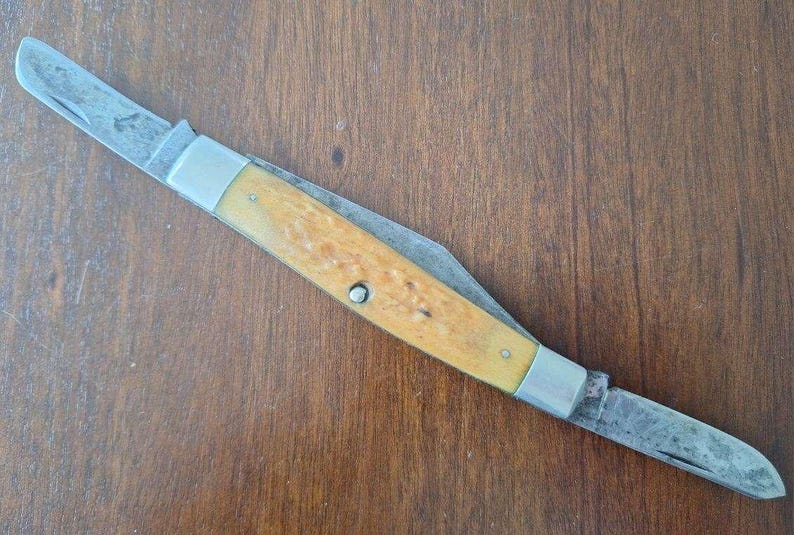 Puede incluir: Una navaja de bolsillo vintage con dos hojas. La navaja presenta un mango de madera de color claro con refuerzos y remaches met&aacute;licos. Las hojas son de acero y muestran signos de desgaste.