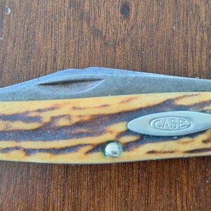 Puede incluir: Una navaja de bolsillo Case con una hoja parcialmente abierta. El mango tiene un patr&oacute;n amarillo y marr&oacute;n, con extremos de metal plateado y un emblema ovalado plateado que dice "CASE". El cuchillo est&aacute; sobre una superficie de madera.