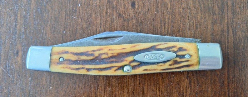 Puede incluir: Una navaja de bolsillo Case con una hoja parcialmente abierta. El mango tiene un patr&oacute;n amarillo y marr&oacute;n, con extremos de metal plateado y un emblema ovalado plateado que dice "CASE". El cuchillo est&aacute; sobre una superficie de madera.