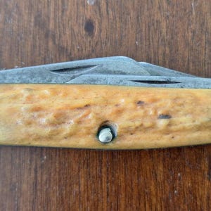 Puede incluir: Una navaja de bolsillo con mango marr&oacute;n claro y detalles met&aacute;licos plateados. La hoja est&aacute; parcialmente abierta, revelando una hoja plateada. El mango tiene un aspecto texturizado y est&aacute; asegurado con un remache plateado.