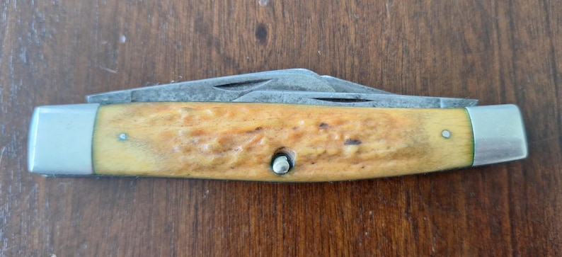 Puede incluir: Una navaja de bolsillo con mango marr&oacute;n claro y detalles met&aacute;licos plateados. La hoja est&aacute; parcialmente abierta, revelando una hoja plateada. El mango tiene un aspecto texturizado y est&aacute; asegurado con un remache plateado.